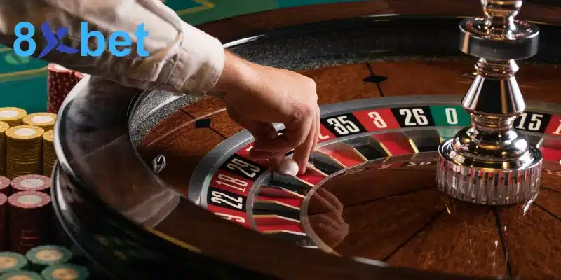 Tay dealer đặt quả bóng trắng vào vòng quay roulette 8xbet bằng gỗ, cạnh các chồng chip cược