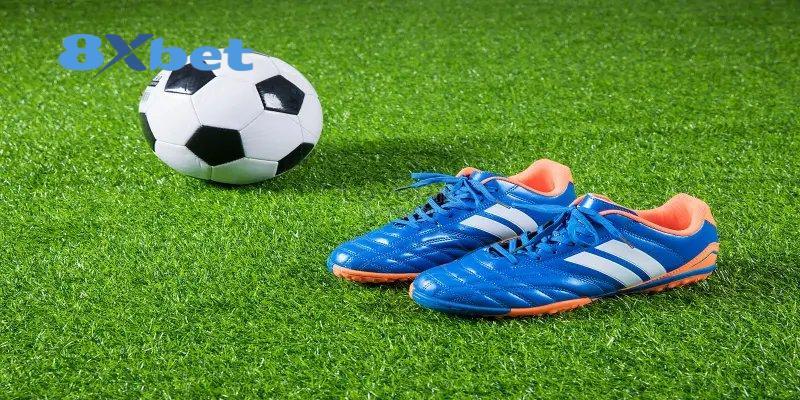 Đôi giày đá bóng xanh cam cạnh trái bóng trên sân cỏ, gợi nhắc danh hiệu vua phá lưới world cup